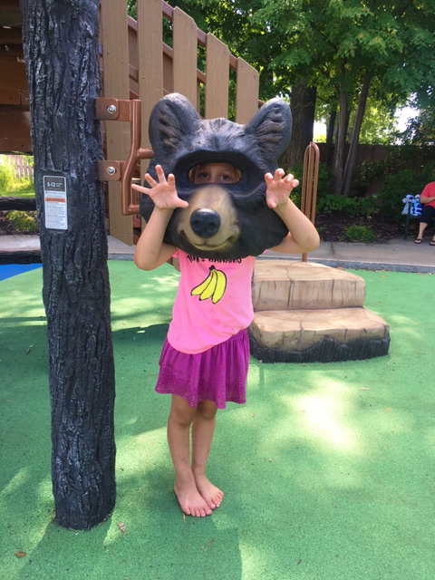 A little girl at a playground structure behind an animal face of a bear, known as a fuzzy face.
