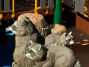 A deck access climber made up of three raccoons and two tree stumps next to a large playground structure